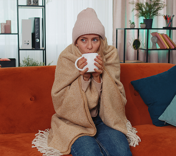Kamer bewoonster op een bank, ingepakt in een deken en muts, drinkt warme drank tegen de kou.