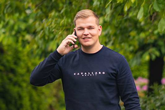 Jan-Thijs staat buiten in een groene omgeving en belt met zijn mobiel, hij draagt een donkerblauwe trui.