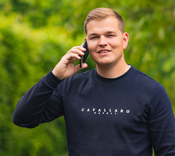 Jan-Thijs staat buiten in een groene omgeving en belt met zijn mobiel, hij draagt een donkerblauwe trui.