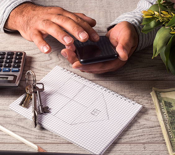 Hand met smartphone naast een rekenmachine, sleutelbos en schets van een huis op ruitjespapier.