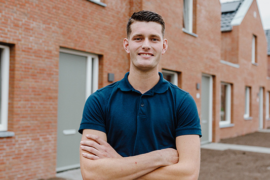 Sven staat met gekruiste armen voor een rij nieuwbouwhuizen en kijkt lachend in de camera.