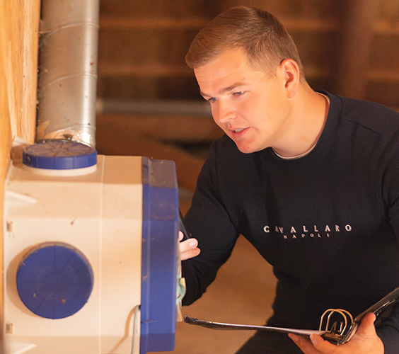 Jan-Thijs inspecteert een ventilatie-unit in een technische ruimte, met een clipboard in zijn hand.